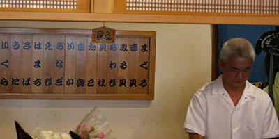 Sushi chef with wooden signs behind him in Japanese listing the seafoods available.